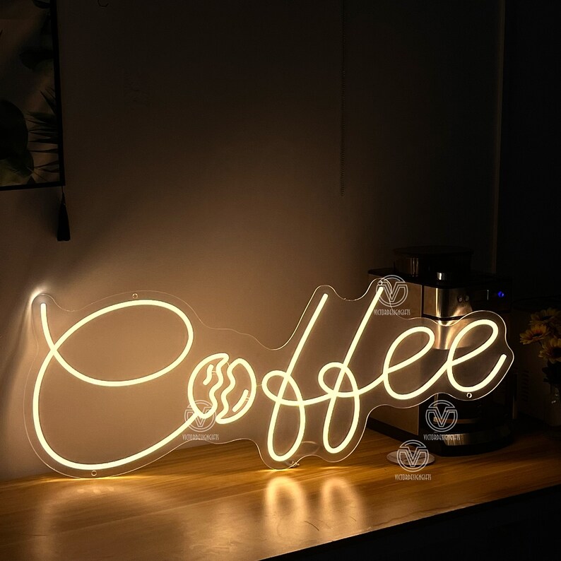 May include: White neon sign with the word "Coffee" in a cursive font. The sign is illuminated and has a coffee bean graphic next to the "C".