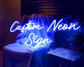 May include: A blue neon sign with the words "Custom Neon Sign" in a cursive font. The sign is illuminated against a dark background, resting on a chair draped with a blue blanket. The sign is a decorative item.