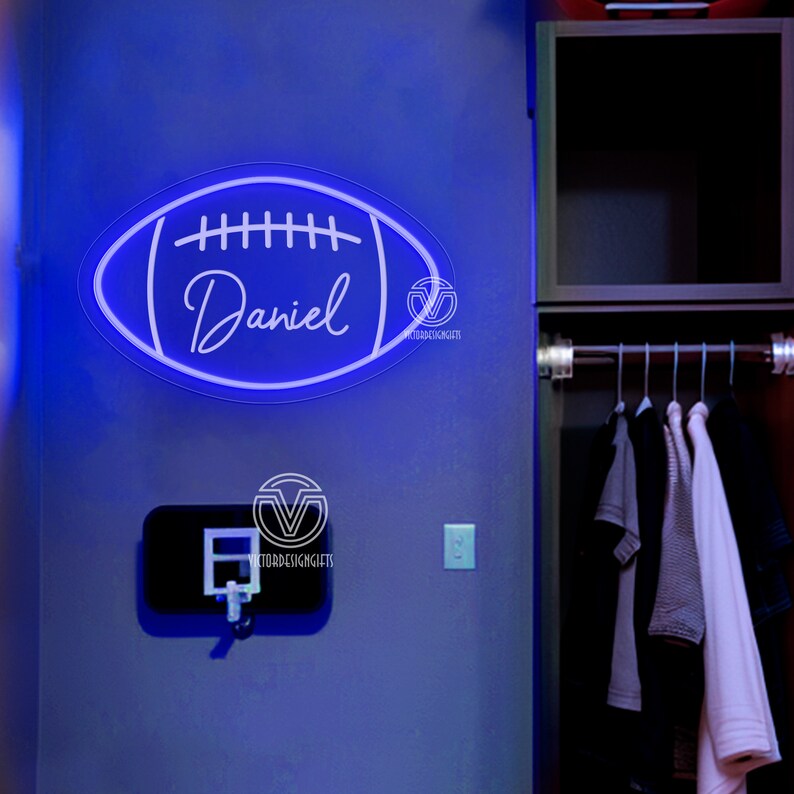 May include: A white neon sign shaped like a football with the name "Daniel" inside. The sign is lit up with a blue glow and is mounted on a grey wall.