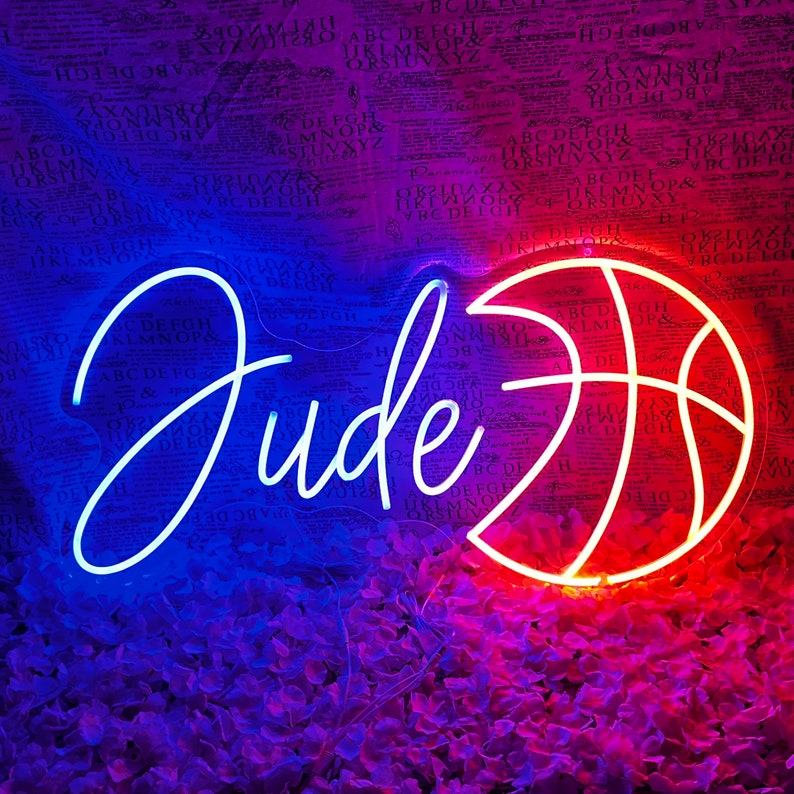 May include: Neon sign featuring the name "Jude" in white script and a basketball in orange and red. The sign is illuminated against a blurred background of a newspaper.