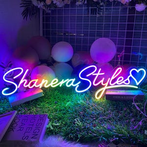 May include: A neon sign with the text "Shanera Styles" in a cursive font, with a heart symbol. The sign glows with a gradient of colors, including blue, yellow, and green. The sign is placed on a bed of green grass.