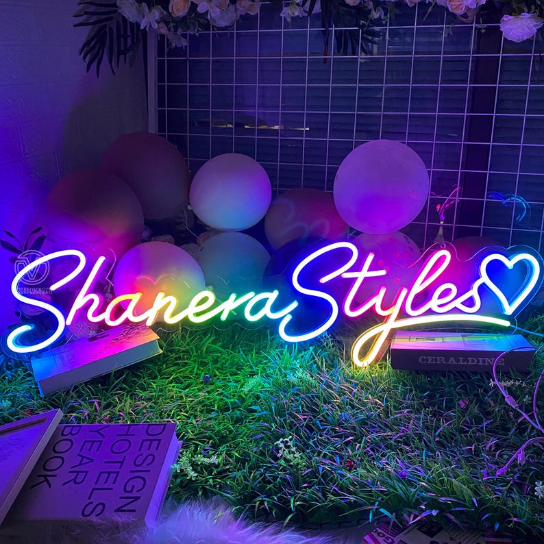 May include: A neon sign with the text "Shanera Styles" in a cursive font, with a heart symbol. The sign glows with a gradient of colors, including blue, yellow, and green. The sign is placed on a bed of green grass.