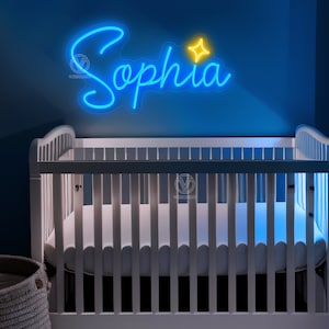K&ouml;nnte beinhalten: Ein individuelles Neonschild mit dem Namen Sophia in leuchtend blauer Schreibschrift, erg&auml;nzt durch ein kleines gelbes vierzackiges Sternsymbol. Das Schild ist auf einer transparenten Acrylplatte montiert und h&auml;ngt &uuml;ber einem wei&szlig;en Babybett.