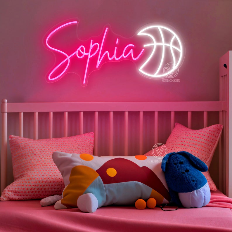 May include: A pink neon sign with the name "Sophia" and a white neon basketball sign. The signs are hanging on a pink wall above a white cot with pink bedding and colourful pillows.