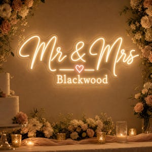 May include: A warm yellow neon sign displaying the text Mr & Mrs with a small heart icon above the word Blackwood. The sign is mounted on a wall and surrounded by floral arrangements, candles, and a tiered cake on the left side.