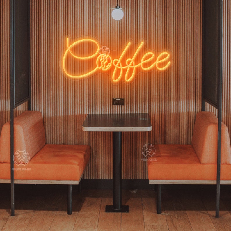 May include: A neon sign with the word "Coffee" in a cursive font with a coffee cup and bean design. The sign is mounted on a wood panel wall with two orange booths and a black table in front of it.