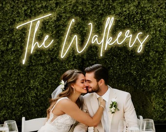 May include: A wedding photograph showing a couple embracing at a table. A neon sign above them reads "The Walkers." The bride is in a white dress and veil, and the groom wears a cream suit. Champagne glasses and a notebook are on the table.