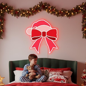 Op de afbeelding: Een rode neon kerststrik en kerstmuts lichtarmatuur is aan een lichtroze muur bevestigd. Een jongen in gestreepte pyjama zit op een bed met een teddybeer. Een kerstslinger met lichtjes en ornamenten hangt erboven.