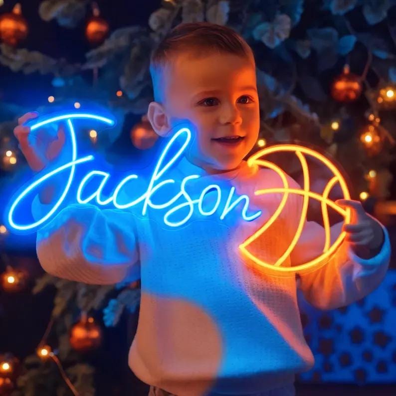 May include: A neon sign in blue script spelling "Jackson" and a basketball in orange. The sign is held by a child in a white sweater. The background is a Christmas tree with lights.