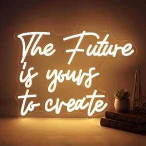 May include: A glowing white neon sign with the words "The Future is yours to create" in a cursive font. The sign is mounted on a clear backing. A small potted succulent and books are visible in the background.