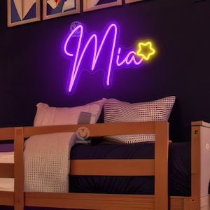K&ouml;nnte beinhalten: Ein individuelles Neonschild mit dem Namen Mia in leuchtend violetter Schreibschrift, erg&auml;nzt durch ein kleines gelbes, leuchtendes Sternsymbol. Das Schild ist an einer dunklen Wand &uuml;ber einem Holzbettgestell mit Kissen und Decke montiert.