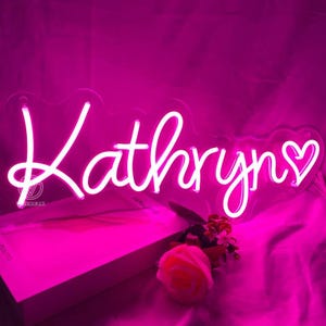 May include: A neon sign in bright pink spells out the name "Kathryns" in a cursive font, with a heart symbol at the end. The sign is illuminated against a pink background. A white box and a pink rose are in the foreground.