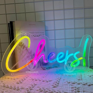 May include: A clear acrylic neon sign with the word "Cheers!" in a rainbow gradient. The sign has a key-like shape and is lit with vibrant colors, perfect for a bar or party setting.
