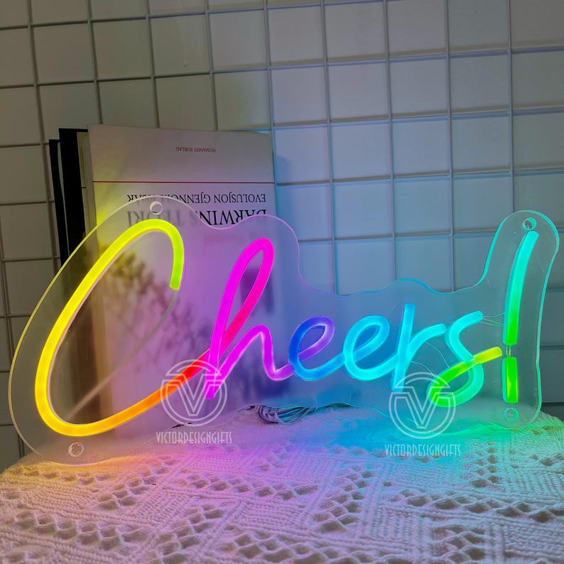 May include: A clear acrylic neon sign with the word "Cheers!" in a rainbow gradient. The sign has a key-like shape and is lit with vibrant colors, perfect for a bar or party setting.