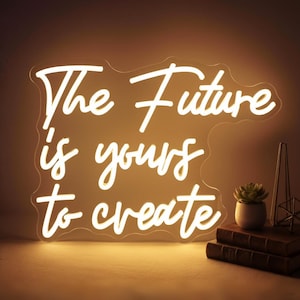 May include: A glowing white neon sign with the words "The Future is yours to create" in a cursive font. The sign is mounted on a clear backing. A small potted succulent and books are visible in the background.