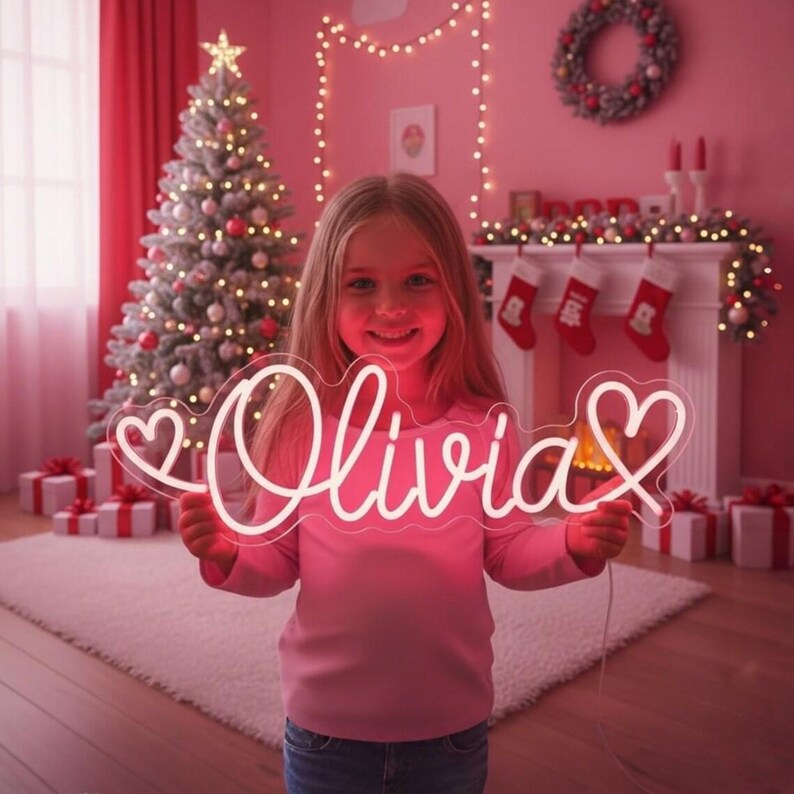 K&ouml;nnte beinhalten: Ein individuelles Neonschild mit dem Namen Olivia in Schreibschrift, flankiert von zwei Herzumrissen. Das Schild leuchtet in hellem Wei&szlig; und wird vor einer festlichen Kulisse mit einem geschm&uuml;ckten Weihnachtsbaum und Weihnachtsstr&uuml;mpfen gehalten.
