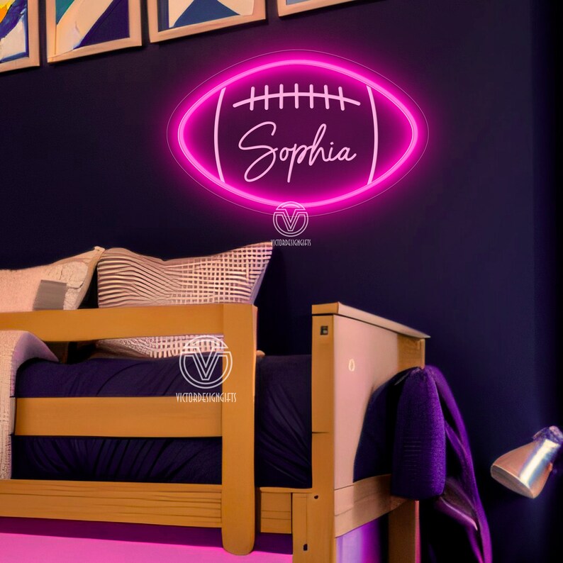 May include: Pink neon sign shaped like a football with the name "Sophia" inside. The sign is hanging on a dark blue wall above a wooden bed.