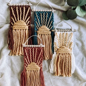 May include: Four woven wall hangings with a sun design. The hangings are made with brown, teal, white, and red yarn. Each hanging has a fringe of yarn at the bottom.