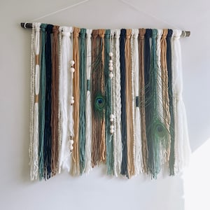 Puede incluir: Un tapiz de pared hecho de hilo en tonos de blanco, marrón, azul verdoso y azul. El hilo está tejido para crear un aspecto texturizado. El tapiz está decorado con cuentas de madera y plumas de pavo real.