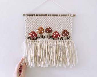 May include: A woven wall hanging with a white background and a fringe border. The hanging features five red and white mushrooms with brown stems.