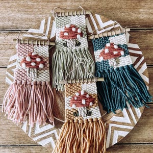 以下が含まれることがあります： キノコのデザインが施された、小さな織りの壁掛けが5つ。それぞれの壁掛けは、異なる色の背景とフリンジが付いています。キノコは白点のある赤色で、白い茎をしています。