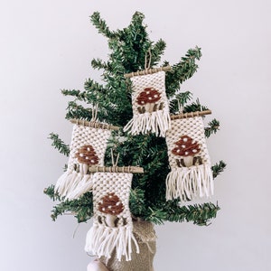 May include: Four woven mushroom ornaments with cream-colored backgrounds and brown mushroom designs are displayed on a small evergreen tree. The ornaments have fringe and are attached to wooden dowels.