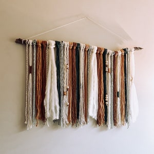 Puede incluir: Un tapiz de pared tejido con una rama de madera natural y varios colores de hilo. Los colores del hilo incluyen blanco, beige, marrón y verde, con cuentas de madera y un acento de cuero. La decoración de la pared está suspendida por una cuerda blanca.