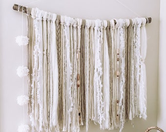 May include: A white and beige macrame wall hanging with pom poms and wooden beads. The hanging is made with a variety of textures and is attached to a natural branch.