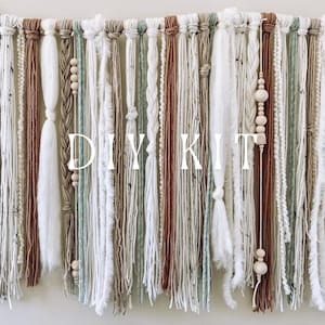 May include: A wall hanging with a DIY KIT theme. The piece features various textures and colors, including white, brown, and green yarn strands. Wooden beads and braided details add visual interest. The text "DIY KIT" is displayed prominently.