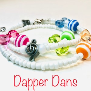 May include: A white beaded bracelet with colorful striped beads, clear beads, and a silver music note charm. The bracelet has a clasp closure. "Dapper Dans" is written in red letters below the bracelet.