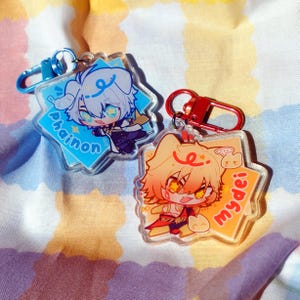 以下が含まれることがあります： アニメスタイルのキャラクターが描かれた、透明なアクリルキーホルダー2つ。1つは青い背景に白髪のキャラクターと「Phainon」という名前が書かれています。もう1つはオレンジ色の背景にキャラクターと「mydei」という名前が書かれています。どちらも金属製の留め金が付いています。