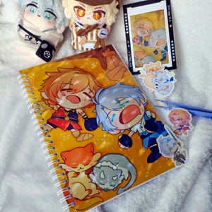 May include: A spiral-bound notebook with a yellow cover featuring cartoon characters and cat illustrations. The image also includes plush dolls, stickers, and a film strip with similar cartoon characters. The items are arranged on a white, fluffy surface.