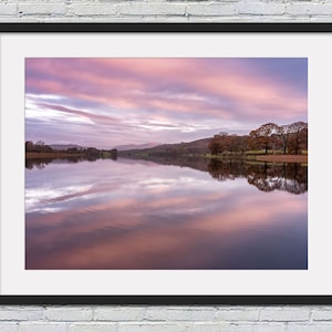 May include: A scenic photograph of a lake with a pink and purple sky reflecting in the water. Trees line the shore of the lake, and mountains are visible in the distance.