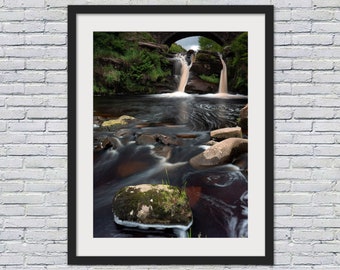 May include: A framed photograph of a waterfall cascading over rocks with a stone bridge in the background. The water is flowing over the rocks and creating a misty effect. The photo is taken from a low angle, giving the viewer a sense of the height of the waterfall.