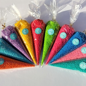 Puede incluir: Un abanico de coloridos conos de fiesta llenos de chispas. Cada cono es de un color diferente, incluyendo naranja, verde azulado, morado, amarillo, rojo, verde, rosa, azul y rosa claro. Cada cono tiene una pegatina con un diseño de copo de nieve.