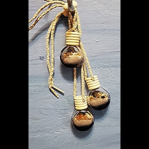 Pode incluir: Três pequenas garrafas de vidro com rolhas de cortiça e detalhes dourados penduradas em uma corda bege. As garrafas estão cheias de uma mistura de materiais dourados e marrons.