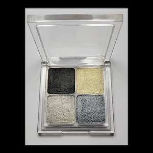 Op de afbeelding: Vierkleurige oogschaduwpalet met zwarte, gouden, zilveren en donkergrijze tinten. De oogschaduws zitten in een transparante plastic compact met een scharnierend deksel. De kleuren zijn sterk gepigmenteerd en hebben een glinsterende finish.