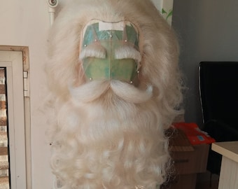 May include: A Santa Claus wig and beard set, featuring long, curly white hair and a matching beard with a prominent mustache. The wig is mounted on a green and white base, and the set is displayed on a black tripod stand.