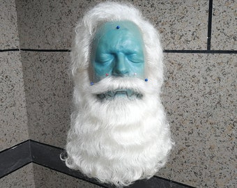 May include: A white wig with a long beard and moustache. The wig is on a mannequin head with a blue face. The mannequin head has several pins in it.