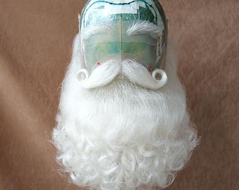 May include: A Santa Claus mask with a white beard and mustache. The beard is full and curly, and the mustache has curled ends. The mask has a green and white head form with red accents.