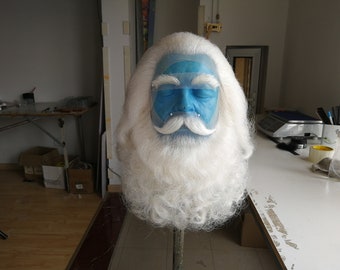 May include: A white beard and mustache wig with a blue face mask. The wig is on a stand and the face mask has a clear plastic shield.