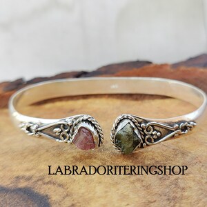 May include: Silver bangle bracelet with two raw gemstone accents. The bracelet is open and has a delicate design. The stones are a pink and green color.