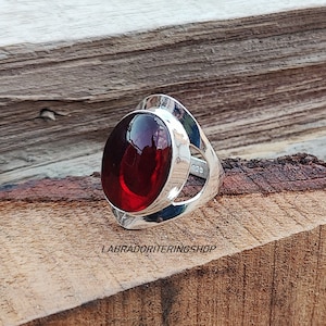 May include: A silver ring with a large, oval-shaped, red gemstone set in a silver bezel. The ring has a unique, open design with a cutout on the side. The ring is marked "925" on the side.