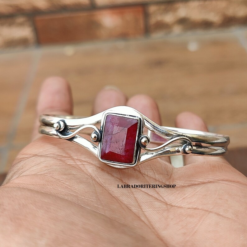 May include: A silver bangle bracelet with a red gemstone set in a square bezel. The bracelet has a delicate, ornate design with two curved bands and small silver beads.