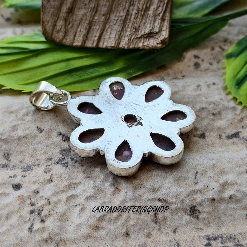 May include: A silver pendant in a flower shape. It has eight petal-shaped insets, each with a dark red gemstone. A small loop is attached for a chain. The text "LABRADORITERINGSHOP" is visible on the pendant.