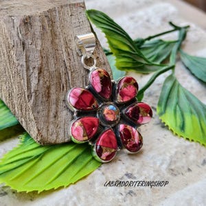 May include: A flower-shaped pendant with a silver bail. The pendant features a cluster of red and pink stones with gold accents. The stones are set in a silver bezel. The pendant is displayed on a wooden block with green leaves.