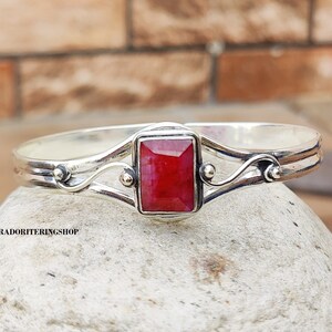 May include: A silver bangle bracelet with a red gemstone set in a square frame. The bracelet has a delicate, ornate design with two curved bands and small decorative beads.