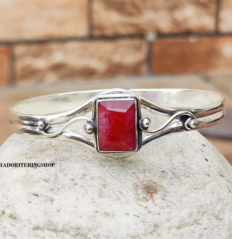 May include: A silver bangle bracelet with a red gemstone set in a square frame. The bracelet has a delicate, ornate design with two curved bands and small decorative beads.