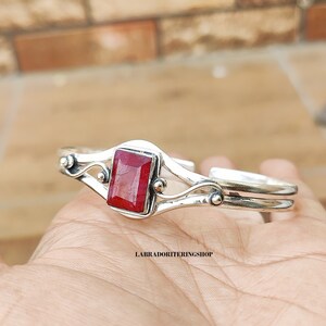 May include: A silver bangle bracelet with a red gemstone set in a square bezel. The bracelet has a delicate, curved design with two bands and small decorative accents.