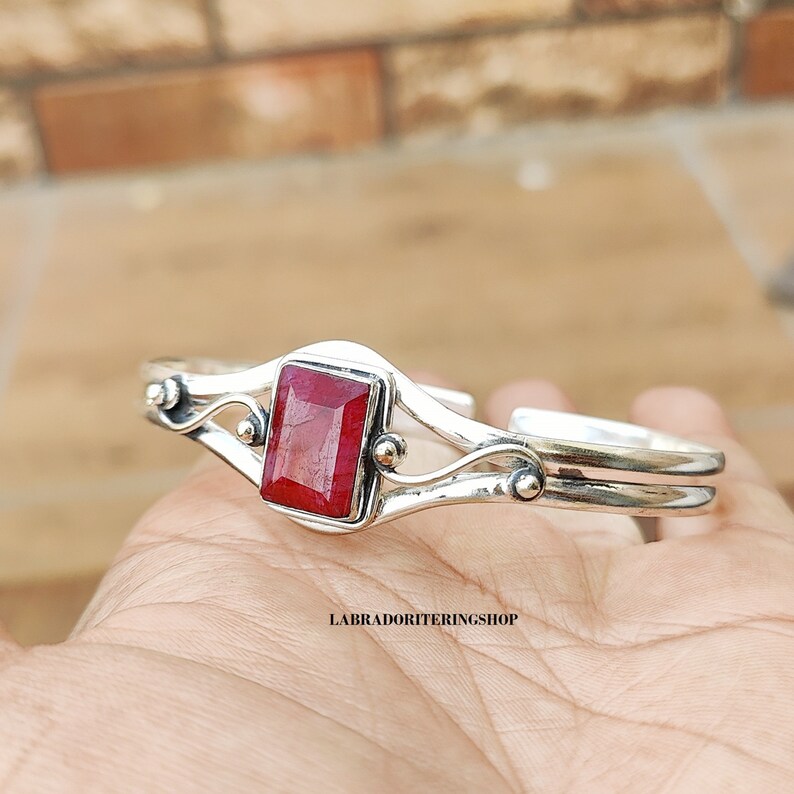May include: A silver bangle bracelet with a red gemstone set in a square bezel. The bracelet has a delicate, curved design with two bands and small decorative accents.